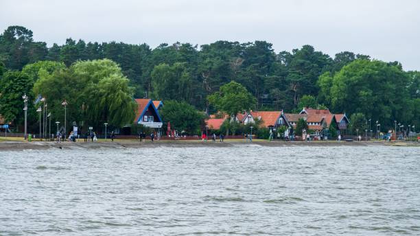 A picturesque lakeside village featuring houses with red roofs, surrounded by lush trees, with people enjoying the waterfront.