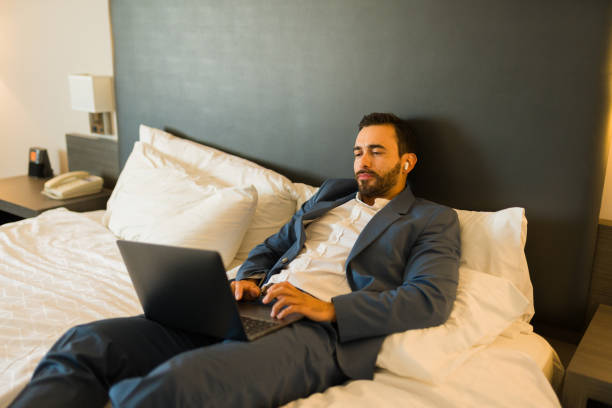 istock Young professional using earbuds and working on a laptop while lounging on a hotel bed 2160128805