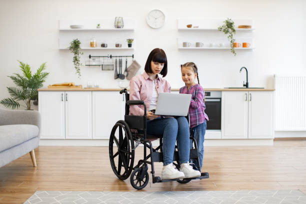 Mother in wheelchair using laptop with daughter in kitchen