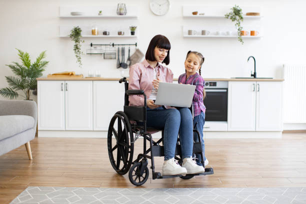 istock Mother in wheelchair using laptop with daughter in kitchen 2160132176