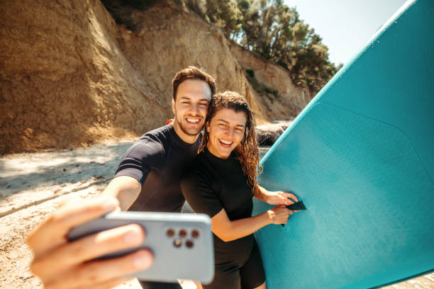 Young beautiful couple taking selfie on the beach, they are holding paddleboard
