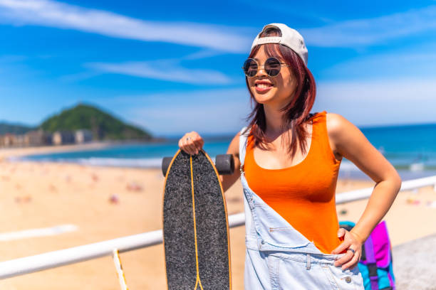 Three quarter length portrait of a chinese cool young woman with skateboard smiling standing next to the beach