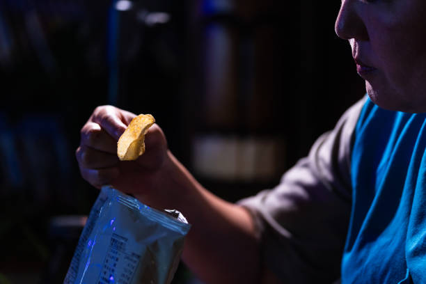 Close up of a woman snacking on potato chips at night while watching a movie in the dark.