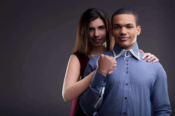Young couple in their 20s or 30s smiling happily, holding arms, exuding love and togetherness against a grey background in a studio setting