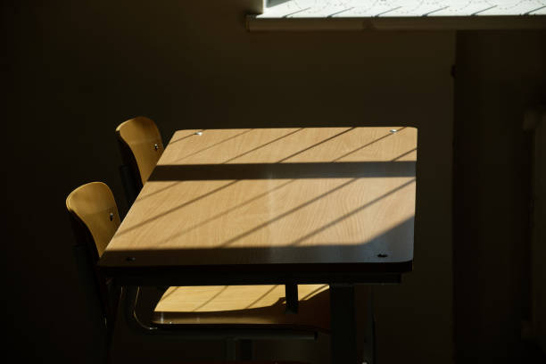Empty wooden desk and chairs in a quiet, dimly lit classroom. Sunlight creates a pattern through the window, highlighting the natural wood texture.