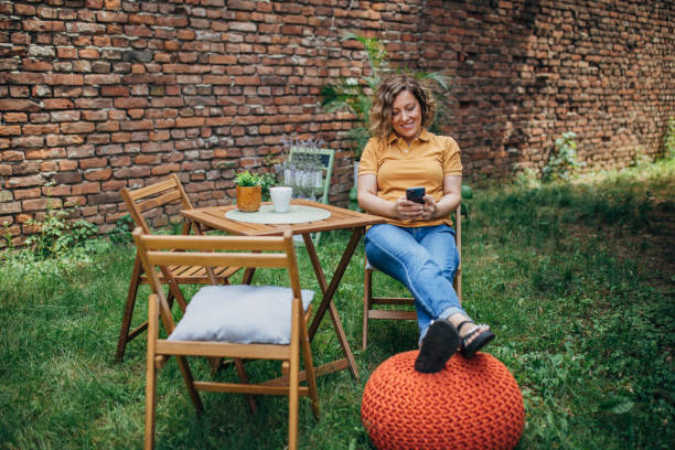 A woman is sitting in a garden on a red stool with her legs crossed, using her smartphone, next to a wooden table set against a brick wall.