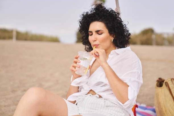 istock A woman is sitting on the beach, drinking a beverage from a plastic cup 2160233090