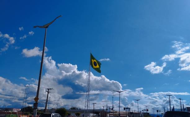 Brazilian flag raised on a public road, fluttering in a blue sky with clouds.