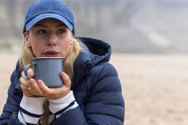 Woman enjoying a misty morning at the coast. She has been for a cold water dip in the ocean and is wrapped up warm, drinking from a mug by the beach.