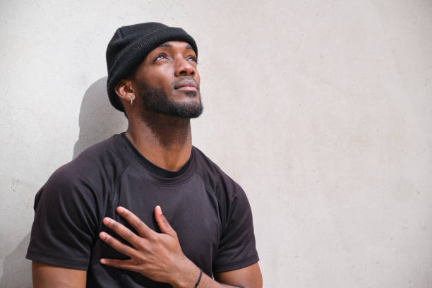 Young man wearing beanie looking up and placing hand over heart Young black man standing with hand over heart while looking up with hope mid adult men stock pictures, royalty-free photos & images