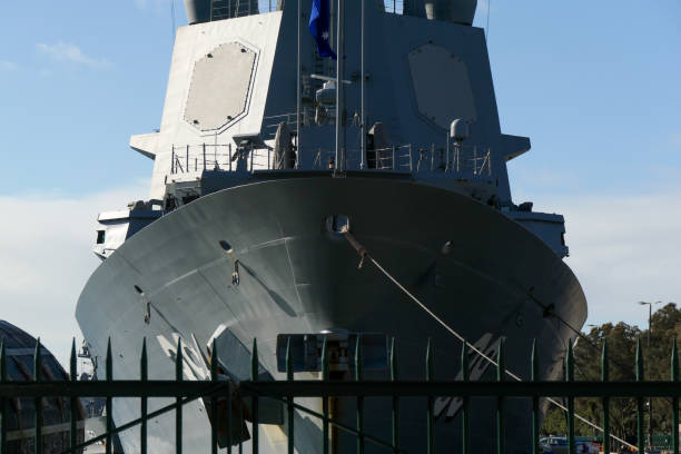 The bow and superstructure of HMAS Hobart, DDG-39, a Hobart Class destroyer of the Royal Australian Navy docked at Garden Island naval base in Sydney Harbour. On the left is a buoy used to allow ships to dock next to each other. In the left background is HMAS Warramunga, FFH-152, an Anzac Class frigate. This image was taken on a cold and sunny afternoon on 1 July 2024