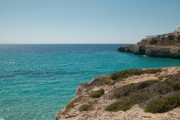 Turquoise mediterranean sea surrounding a rocky cliff on a sunny day, with a luxury resort in the background