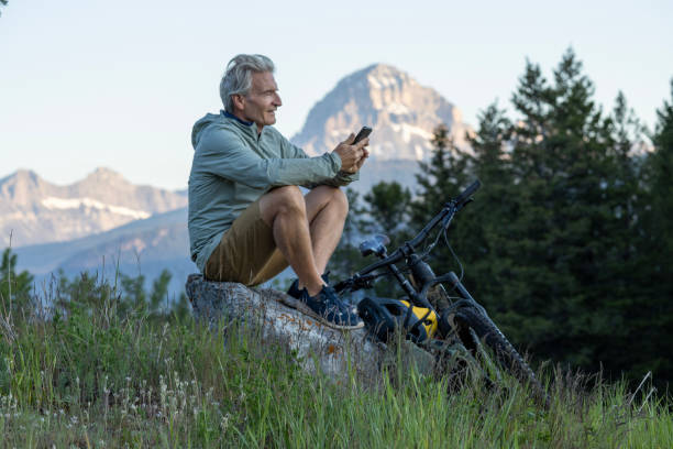 Mountain biker uses mobile phone in meadow Mountain biker uses mobile phone in meadow below snowcapped mountain, Crowsnest Mountain man 60s bicycle stock pictures, royalty-free photos & images