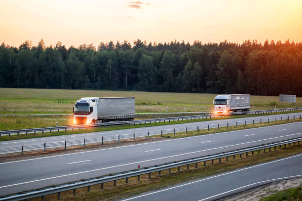A column of truck tractors with semi-trailers and headlights on are transporting cargo along the highway against the backdrop of sunset and summer. Concept of working as a truck driver, remuneration, logistics una columna de camiones tractores con semirremolques y faros encendidos transportan carga a lo largo de la carretera con el atardecer y el verano como telón de fondo. concepto de trabajo como conductor de camión, remuneración. copiar espacio para texto