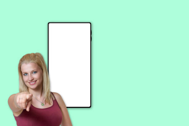 Smiling attractive young blond woman in sportswear is finger pointing at the camera. The blank screen  of  smartphone is ready for your text