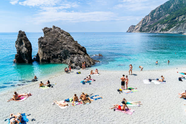 Scenic beach in Monterosso, Cinque Terre, featuring distinctive rock formations and sunbathers enjoying the clear blue water. The sandy shore is dotted with people relaxing on towels. The green hills and rocky cliffs in the background enhance the natural beauty of the coastal landscape.