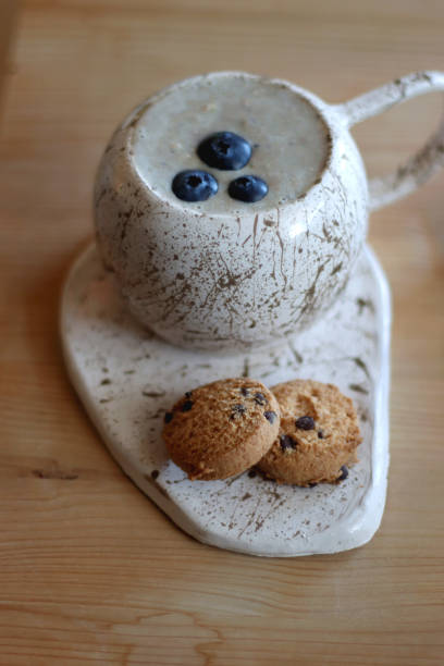 Cup of blueberry and banana smoothie, chocolate chip cookies, book, pen and reading glasses on the table. Colorful bookshelf in the background. Selective focus.