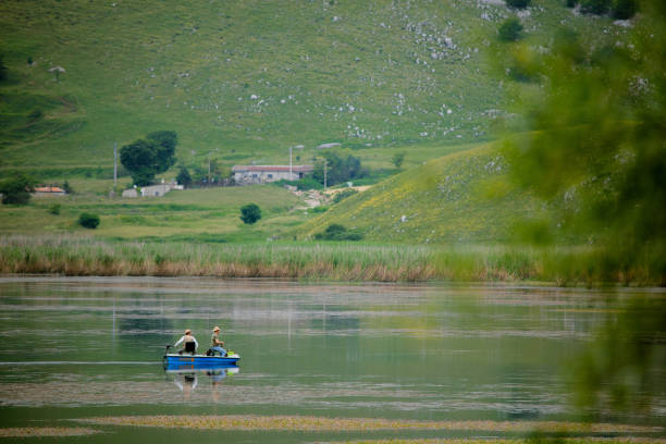 A bright yellow fishing rod extends into the frame, its tip and line in crisp focus against the vibrant, blurred backdrop of a shimmering alpine lake. The turquoise water glows with patches of green and blue, bordered by lush grasses and smooth shoreline rocks. Towering evergreen trees line the distant shore under a brilliant blue sky, creating a tranquil scene perfect for anglers and nature lovers alike.