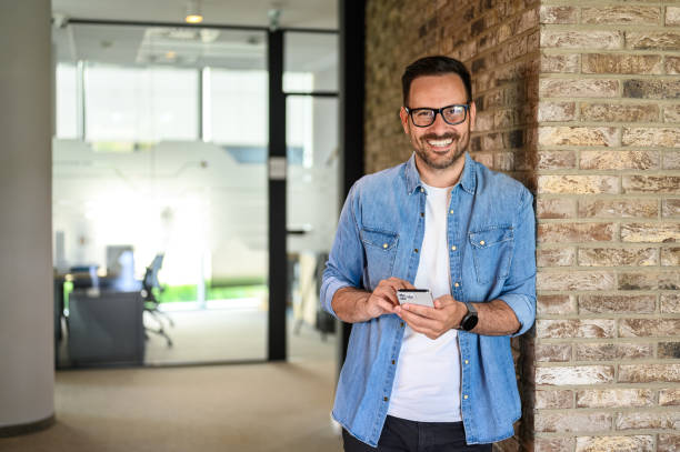 istock Portrait of happy young man developing mobile phone applications while leaning on brick wall in office 2160288047