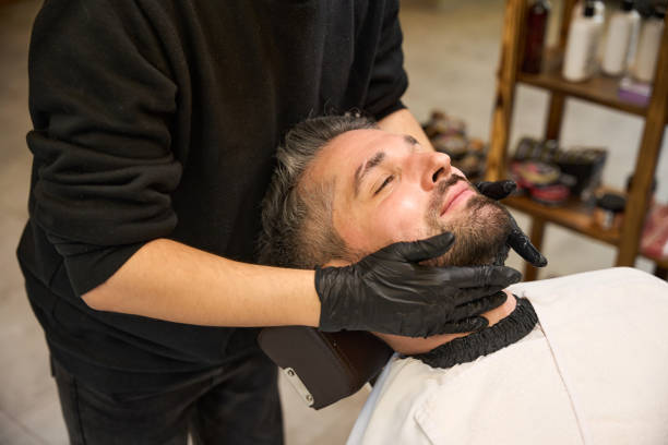 Tender barber in protective gloves applying the beard balm to style and maintain flyaways to male client after trimming and modelling, grooming finish steps