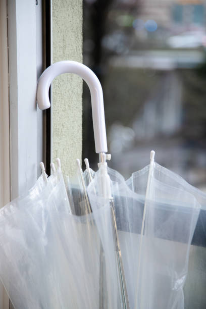 A clear umbrella with a white handle rests against a window, capturing the serene ambiance of a rainy day and the delicate balance of indoor and outdoor elements.