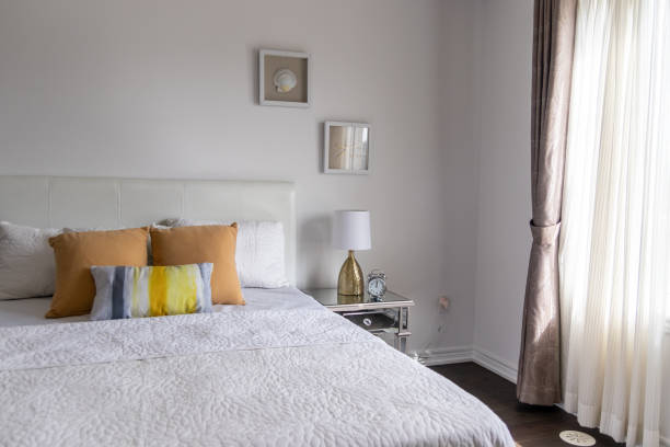 Brightly-lit bedroom with white bedspread, tan and yellow accent pillows, mirrored side table. Taken in Toronto, Canada.