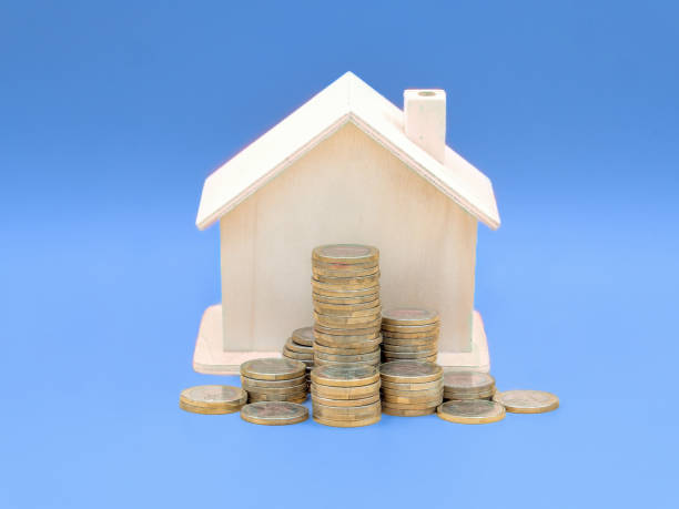 istock Wooden House Model with Stacked Coins on Blue Background 2160355598