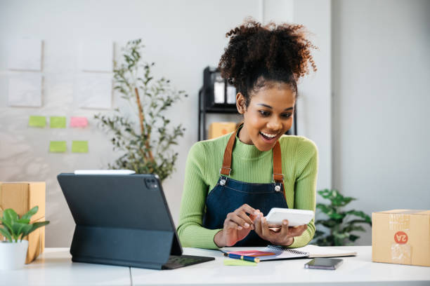 istock African Owner SME Small and Medium Enterprises woman received orders from clients and check profit on calculator preparing products in parcel cardboard box packaging at home ready for delivery 2160368098