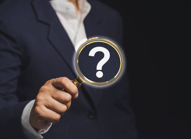 istock Businessman holding a magnifying glass with a white question mark symbol while standing against a black background. 2160377280