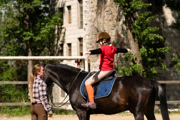 Child rider practices balance on horse guided by instructor. Riding school