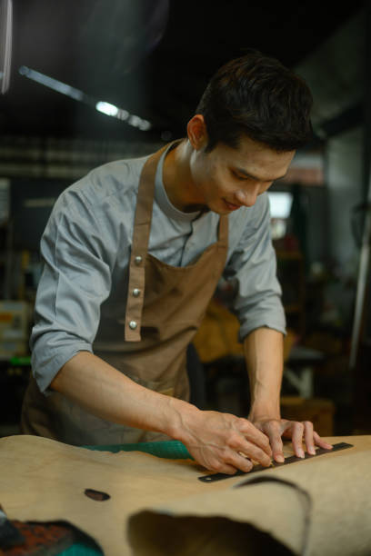 istock Handsome male tailor in apron measuring and cutting brown leather at his workshop 2160400114