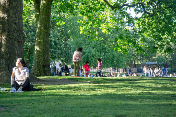 London, UK -  May 21, 2017: People sitting on the grass in St James Gardens in London enjoying the sunshine. A woman with two young children is also pictured.