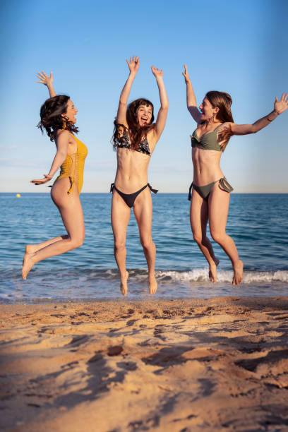 Three young dutch women jump on beach near sea water. A white sea water wave is under their bodies. By jumping so high, they express their enthusiasm, emotion, joy and pleasure. All three of them have beautiful long blond hair. These caucasian teenage girls are on vacation in europe together. They are jumping of joy in a row on the shore with the sea water on the background. They are enjoying their holidays on this sunny day in summer season. The sunshine has colored their skin to a nice brown color.