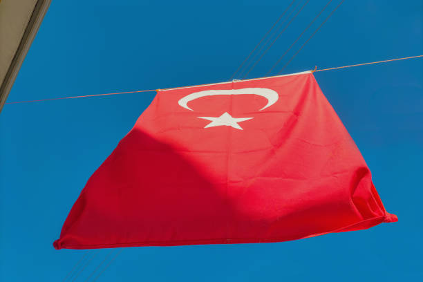 Turkey's red flag with a white crescent moon and a five-pointed star hangs on a rope between houses against a clear blue sky. Symbols of statehood, patriotism, signs, symbolism