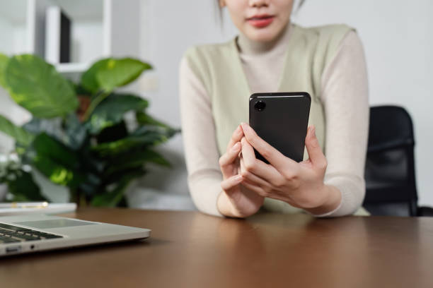 istock Young Professional Woman Working Remotely from Home Office Using Smartphone and Laptop, Green Plant in Background 2160416479