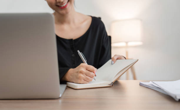 istock Young Woman Studying Online at Home on Laptop, Taking Notes in Notebook, Modern Home Office Setting 2160417210