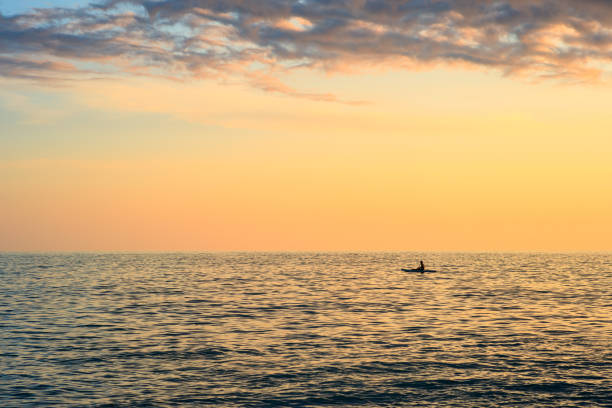 A man is swimming in the ocean at sunset. The sky is a mix of orange and pink hues, creating a serene and peaceful atmosphere