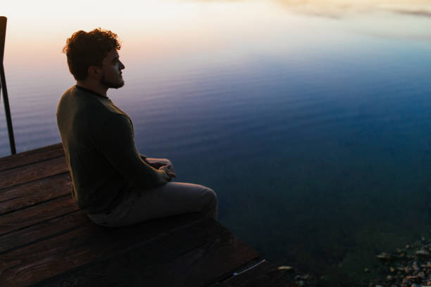 Young man sitting on a pier on a lake. The atmosphere is calm and tranquil, he looks meditative and relaxed.