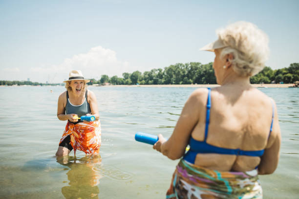 Happy senior woman having fun on holiday, standing in water and playing with squirt gun.