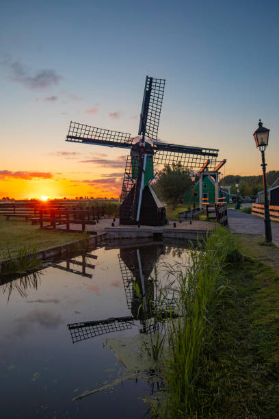 A breathtaking rural scene at the famous tourist site Zaanse Schans in Zaandam, Netherlands. This image captures the iconic Dutch windmills at sunset, with a dramatic sky painting the backdrop in vibrant hues. The windmills, with their historical significance, stand tall and majestic, reflecting the rich cultural heritage of Holland. The tranquil waters of the nearby canal mirror the colorful sky, enhancing the serene atmosphere. The setting sun casts a warm glow, adding a golden touch to the landscape, making it a perfect representation of Dutch rural charm and timeless beauty.