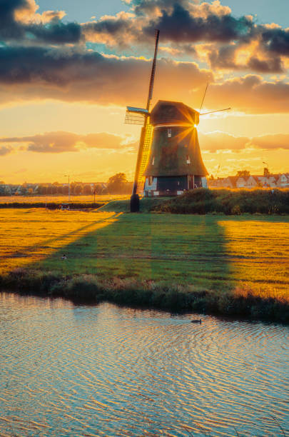Witness the enchanting beauty of a Dutch landscape adorned with vintage windmills at sunset in South Holland. The scene captures old antique windmills standing proudly against a dramatic and colorful sunset sky. As the sunbeam and flare cast a warm glow, the reflection of these historic wooden windmills shimmers in the tranquil canal waters below. This picturesque view of the Dutch polder landscape evokes a sense of timelessness and nostalgia, highlighting the rich cultural heritage and scenic charm of the Netherlands. The combination of natural beauty and historic architecture makes it a captivating destination for photographers and travelers.