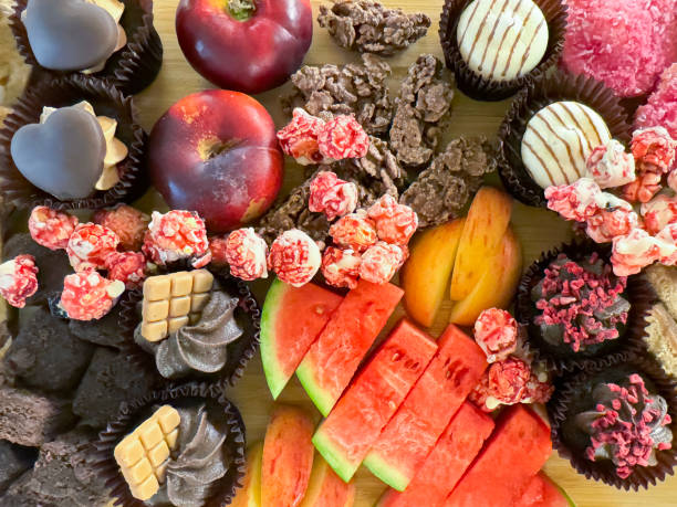 Stock photo showing elevated view of dessert party buffet board covered with cakes, sliced and chopped ingredients including chocolate brownie squares, mini chocolate cupcakes with milk chocolate, toffee and white chocolate butter icing, red apple and watermelon slices, nectarines, caramel shortcake, white chocolate raspberry popcorn, coconut cakes and chocolate crispy squares.
