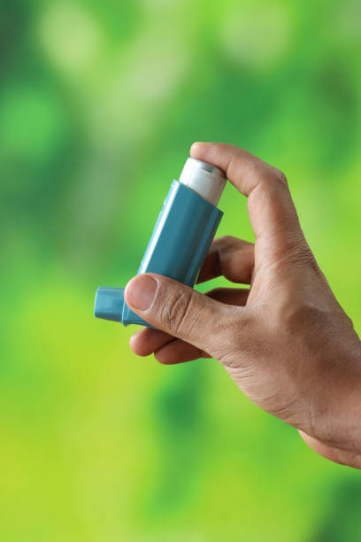 Stock photo showing close-up view of an unrecognisable person holding a metered-dose inhaler used for dispensing medicine to relax and open up airways to the lungs after a bronchospasm caused by exercise or allergy induced asthma. imagen de una persona irreconocible sosteniendo un inhalador para aliviar el asma, el dedo índice presionando el recipiente del inhalador de dosis medida encerrado en un soporte de plástico azul (actuador), la tapa quitada de la boquilla, fondo verde bor