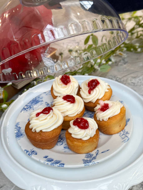 Stock photo showing close-up view of a restaurant cafe counter display of cakes and desserts on cake stands with glass cloches. imagen de cúpula de vidrio de campana de pastel levantada en soporte de pastel que contiene un plato con motivos florales blancos y azules de cupcakes de bizcocho victoria, cupcakes de vainilla cubiertos con remolinos de crema de mantequilla y mermelada d
