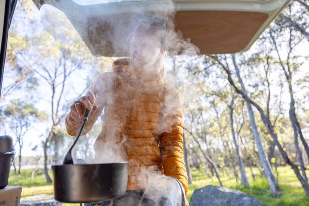 Woman in a yellow jacket stirring a pot with steam rising inside a camper van, with a forest view in the background, highlighting the experience of outdoor cooking and van life.