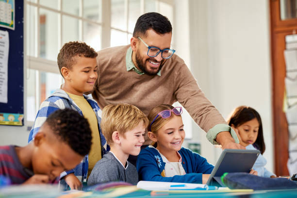 Primary multiethnic pupils working on digital tablet with teacher Cheerful male teacher helping students using digital tablet in classroom during technology lesson. Helpful male professor talking to multiethnic students using digital tablet in classroom. Male teacher supervising a group of elementary children working on computer. schoolboy stock pictures, royalty-free photos & images