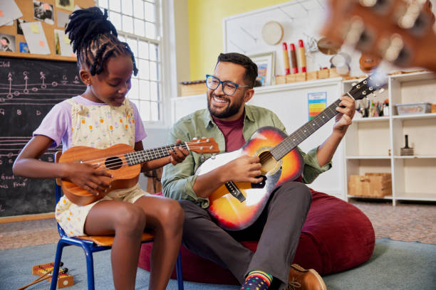 Teacher and students playing guitar together during music lesson Happy male teacher assisting an african kid to play musical instrument in classroom at school. Happy teacher playing acoustic guitar and singing while having music class with girl. Cute little girl learn to play ukulele with male teacher. ukulele stock pictures, royalty-free photos & images