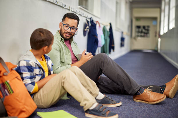 Teacher talking to young sad student Young male psychologist speaking to depressed boy in school while sitting on the floor in school corridor. Man working with a child while discussing about learning problem and bullying. Young and friendly teacher speaking to boy in school hallway. adult schoolboy teacher student stock pictures, royalty-free photos & images