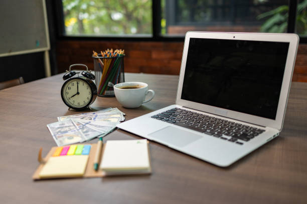 Office workspace featuring a laptop, alarm clock, coffee, colorful stationery, and a pile of hundred-dollar bills.