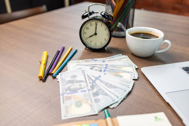 Office workspace featuring a laptop, alarm clock, coffee, colorful stationery, and a pile of hundred-dollar bills.